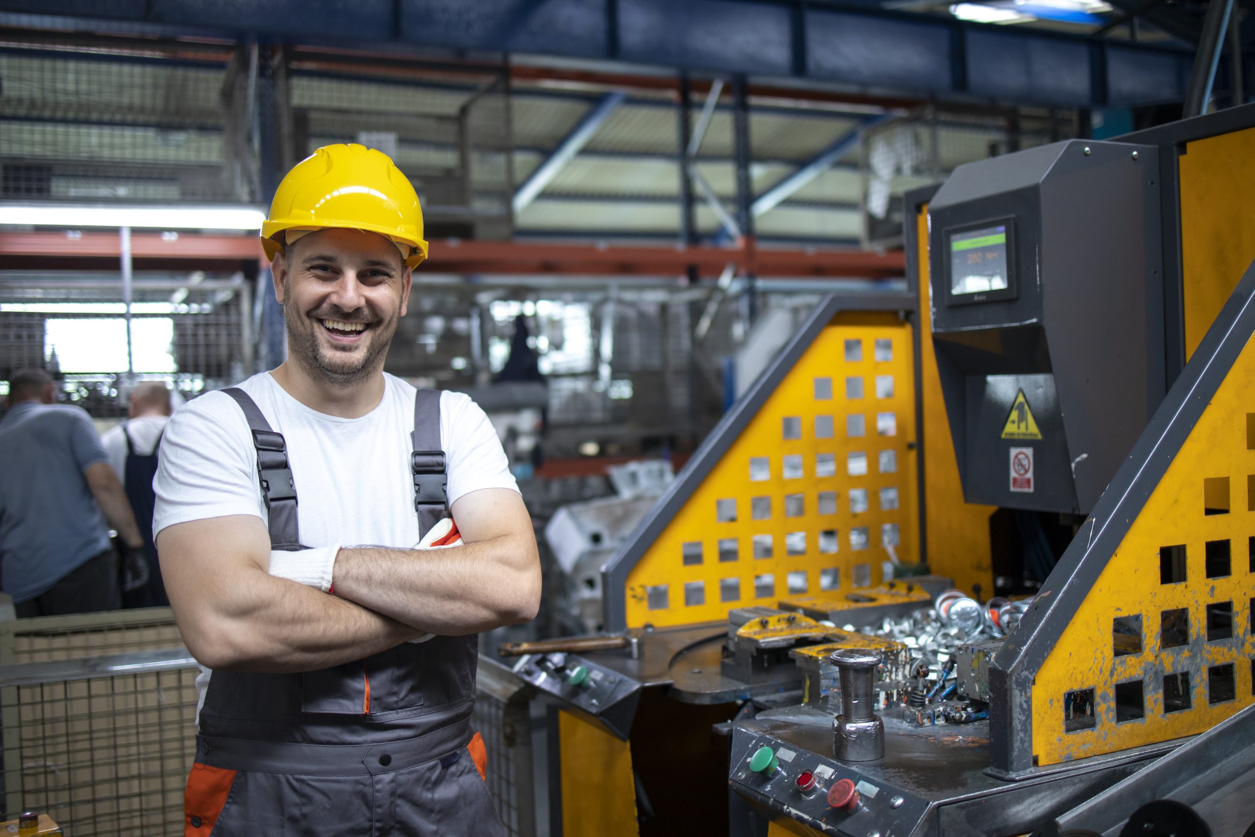 Portrait of factory worker with arms crossed standing by industrial machine in production plant.